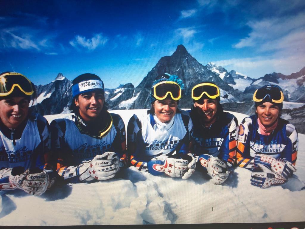 Cinco esquiadoras jóvenes con gafas, apoyadas en la nieve ante las montañas